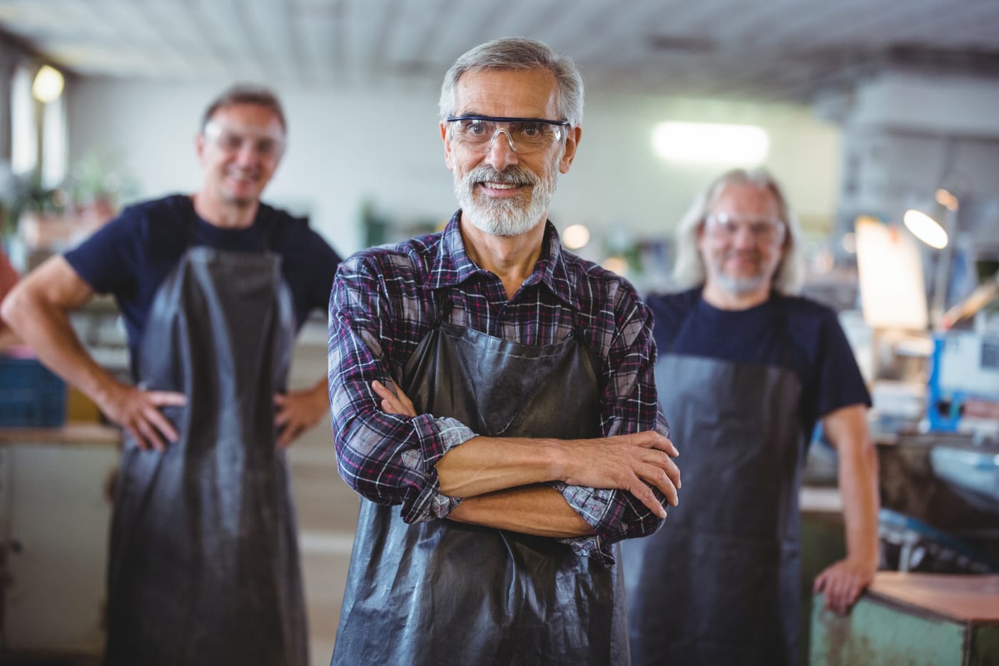Trois artisans souriants en tabliers et lunettes de protection, dont un au premier plan les bras croisés
