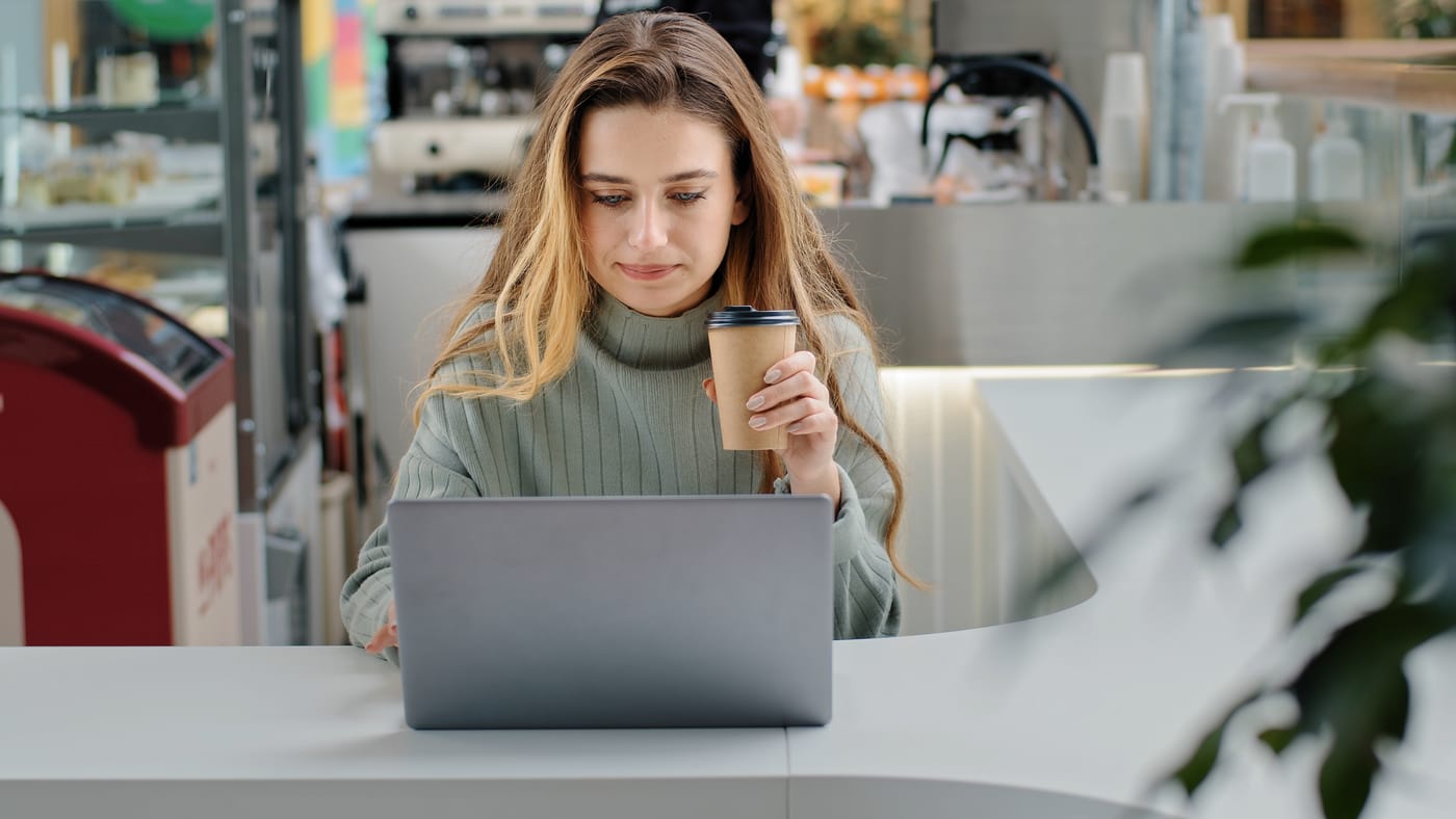 Jeune femme concentrée sur son ordinateur portable, tenant un café, assise à un comptoir dans un commerce